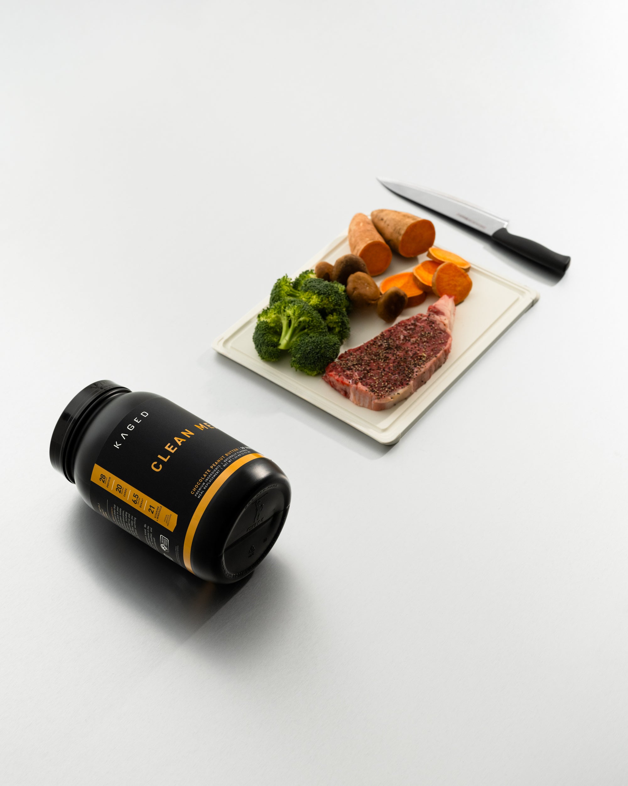 Angled view of KAGED Clean Meal jar with CLEAN MEAL label beside a cutting board with steak and vegetables on gray surface