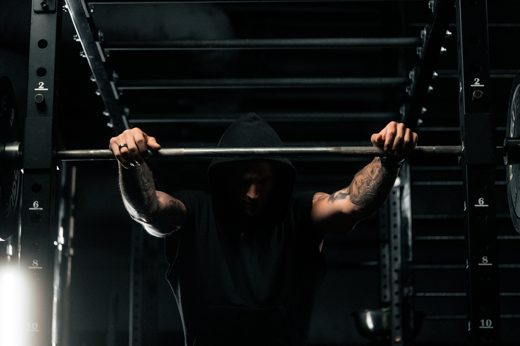 Man in hooded sleeveless top bracing a barbell on a squat rack in a dimly lit gym