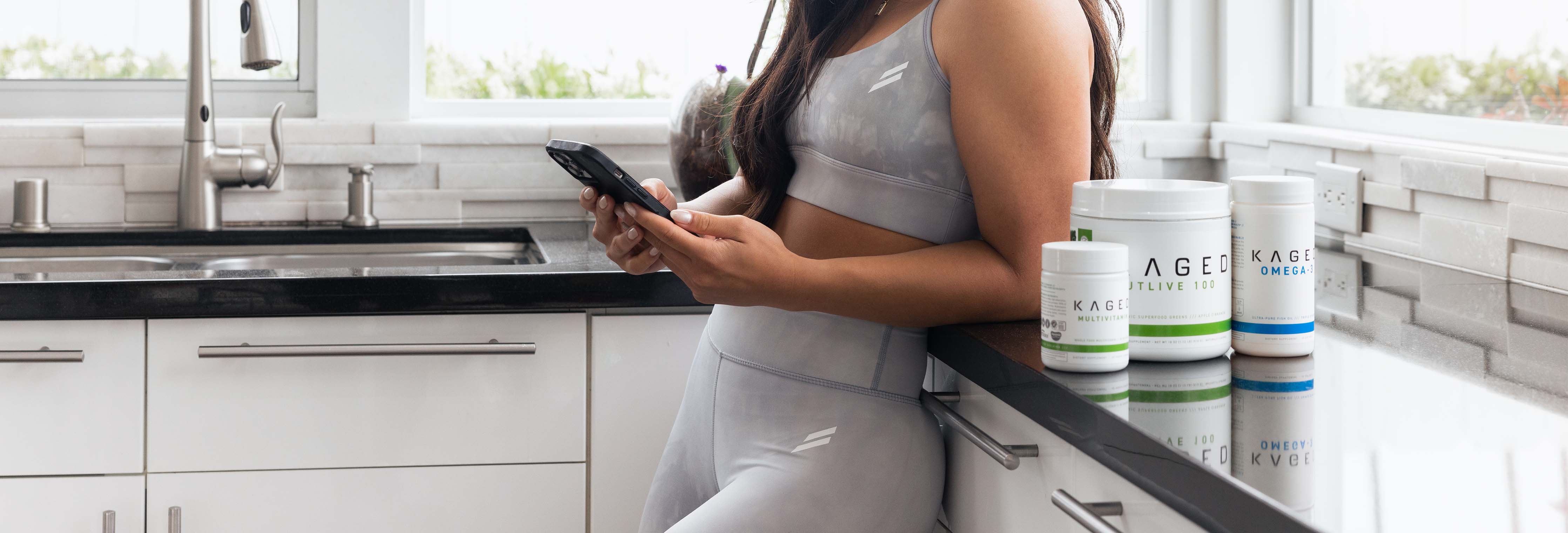 Woman in gray activewear leaning on a kitchen counter checking her phone beside Kaged supplement containers
