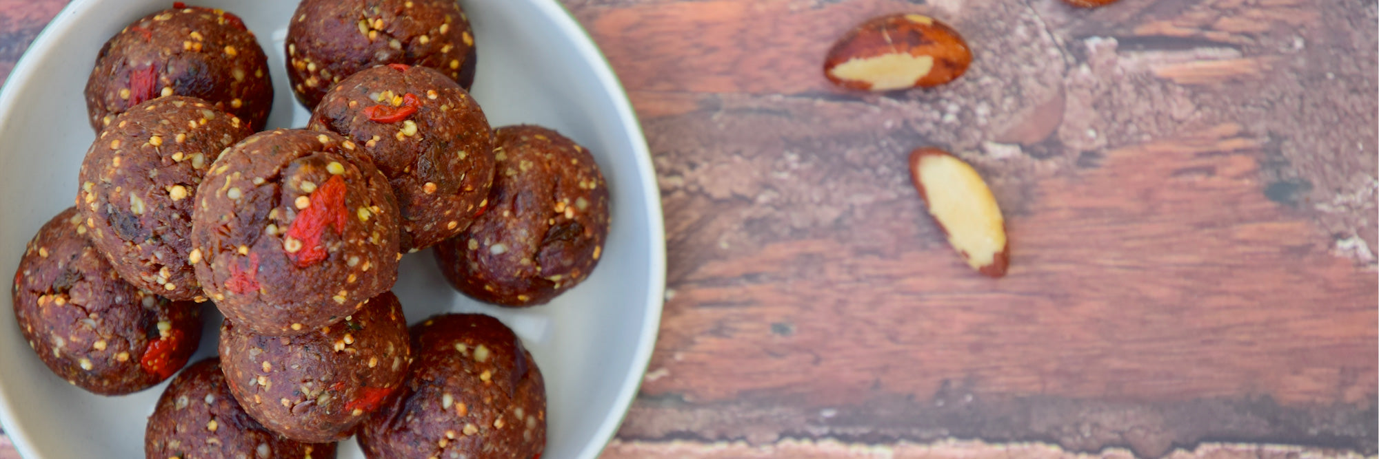 Top-down view of chocolate peanut butter protein balls in a white bowl showing seeds and goji pieces on a wooden surface