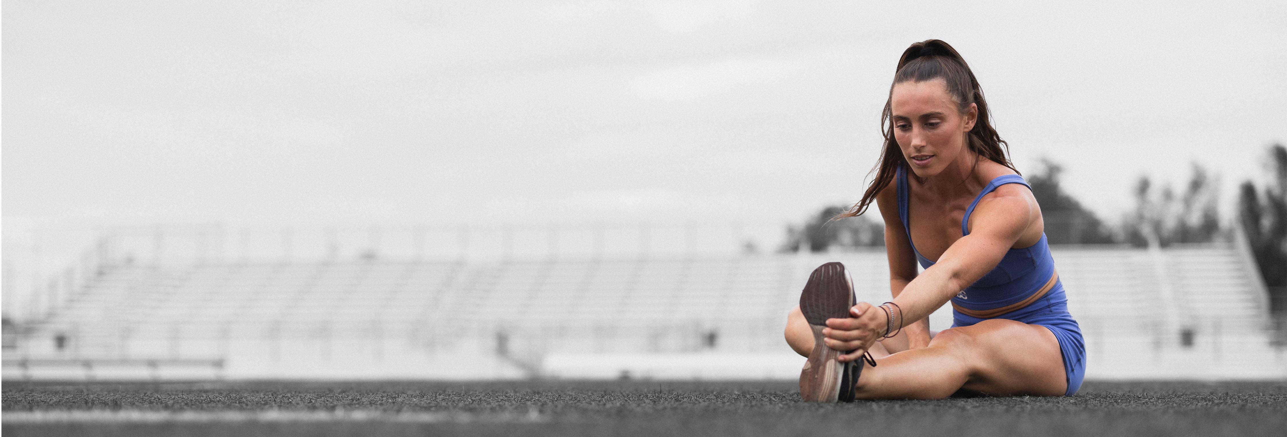 Woman stretching hamstring on outdoor track before a strength training session, empty bleachers in background