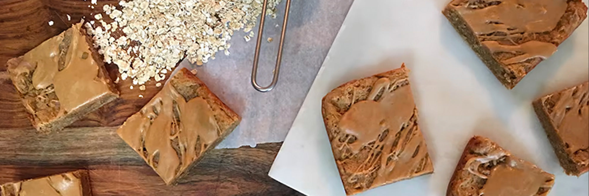 Overhead view of protein peanut butter blondie squares with peanut-butter drizzle and oats on parchment and a wooden board