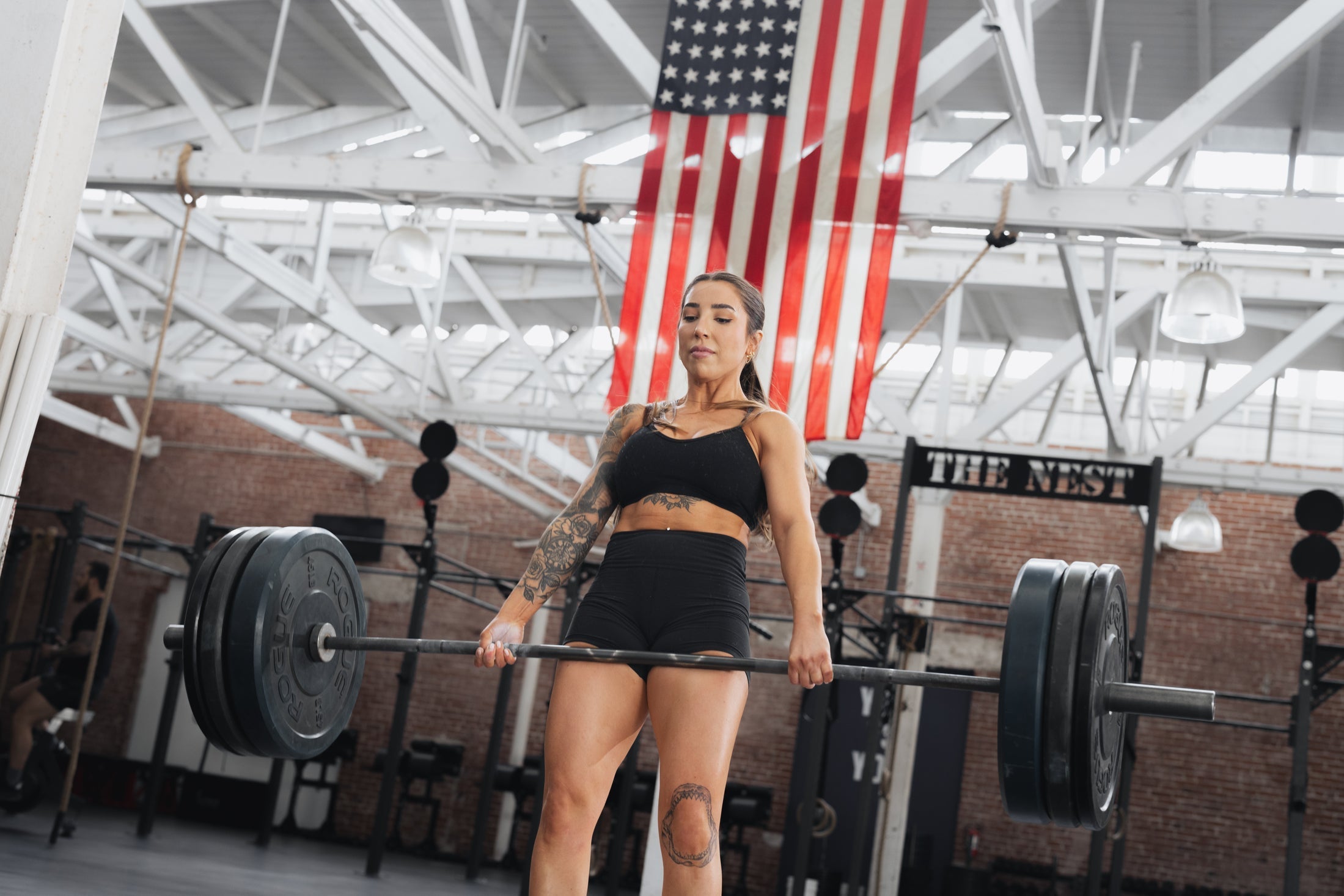 Woman deadlifting a heavily loaded barbell in a brick-walled CrossFit gym with an American flag overhead