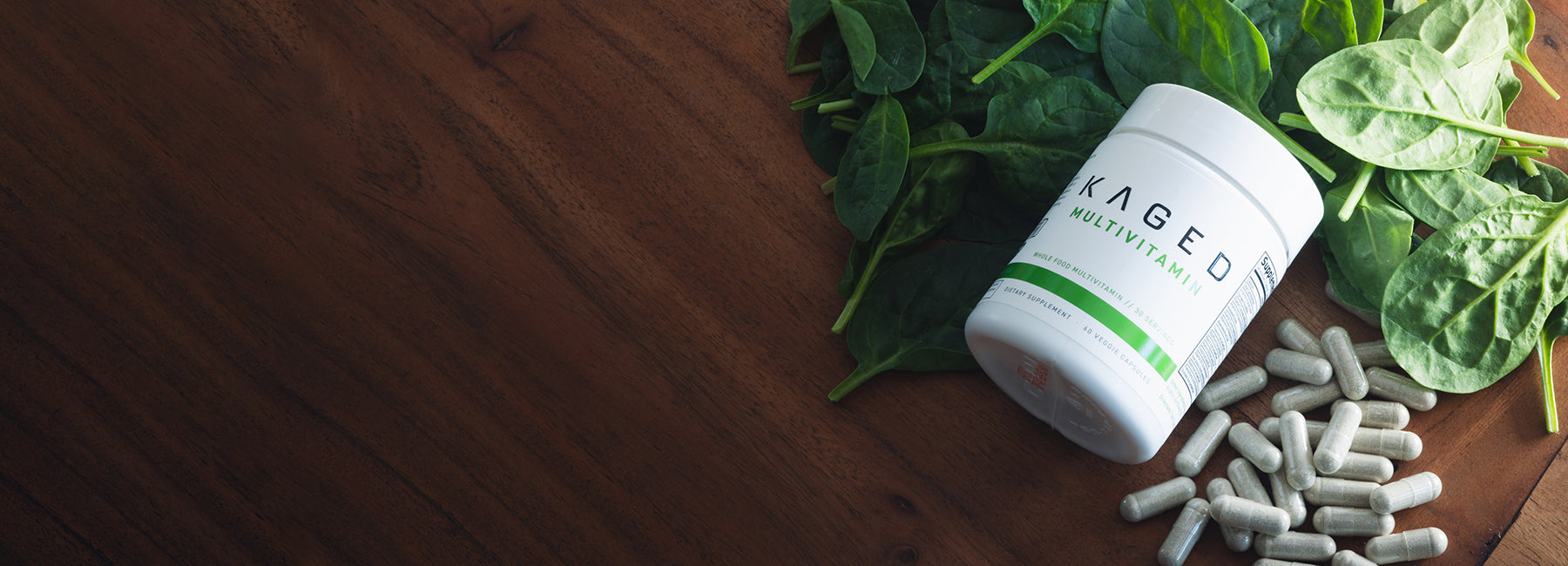 Angled view of KAGED multivitamin bottle with capsules and fresh spinach on a wooden table
