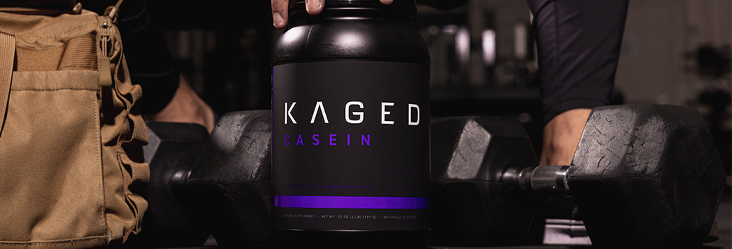 Front view of KAGED Casein tub with purple accent label between dumbbells on a gym floor