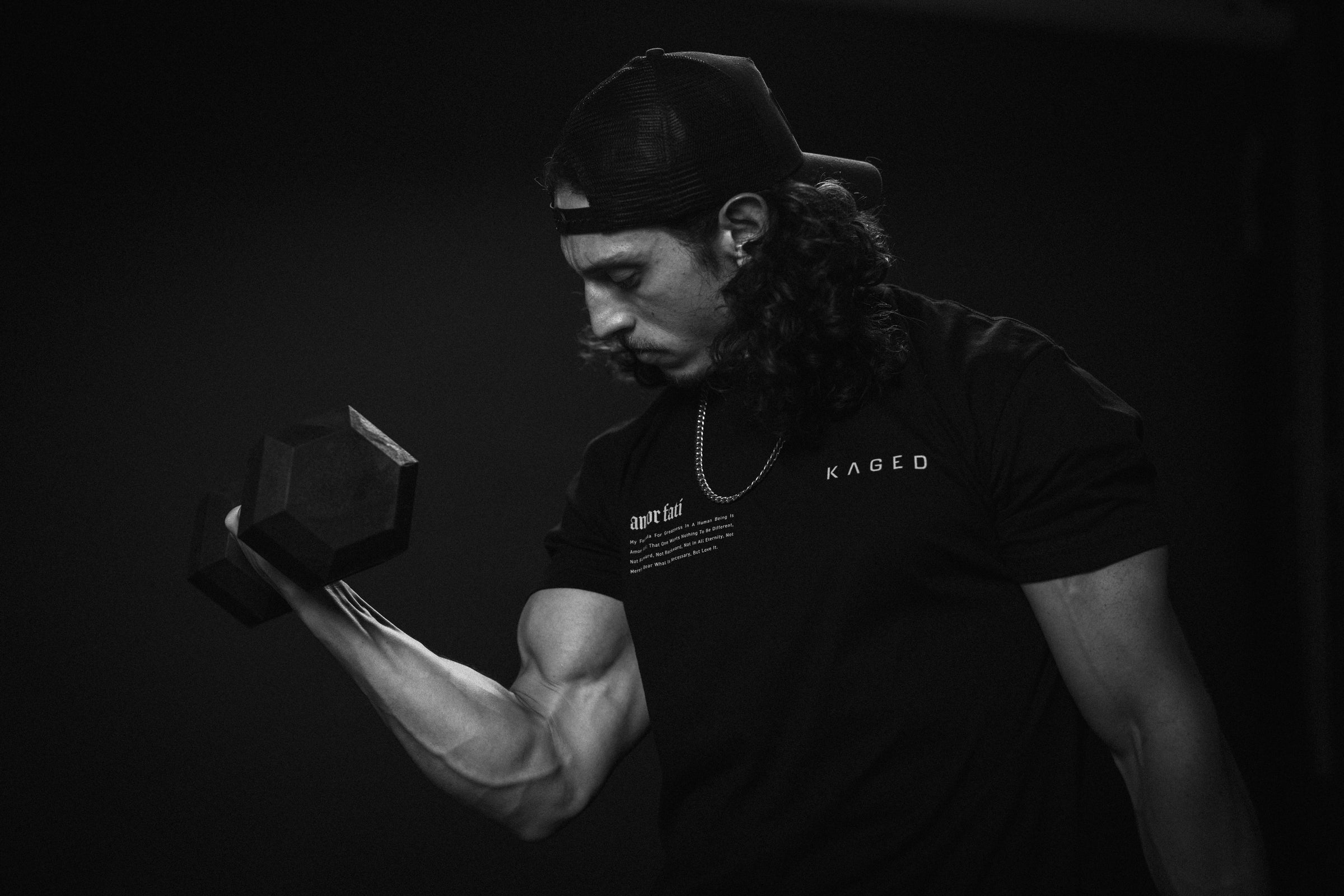 Man performing a dumbbell biceps curl in a dark studio, muscular veiny forearm visible, wearing a KAGED shirt