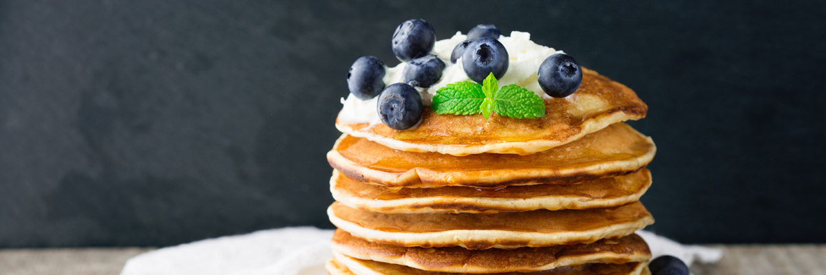 Stack of protein pancakes topped with whipped cream, fresh blueberries, and mint on a dark background