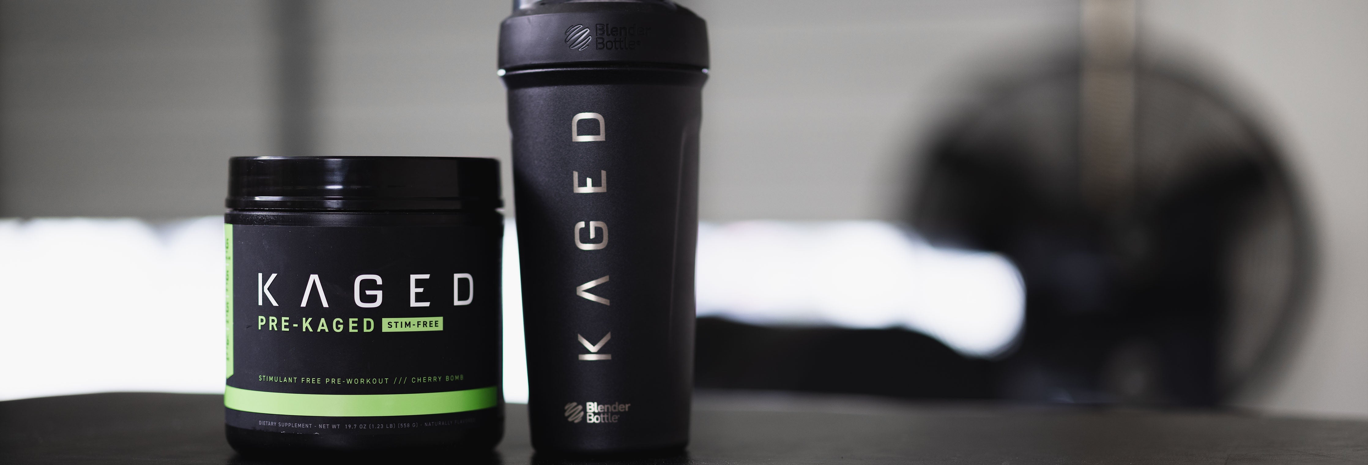Front view of KAGED Pre-Kaged stim-free pre-workout tub beside a KAGED shaker bottle on a gym table with a blurred fan in the background