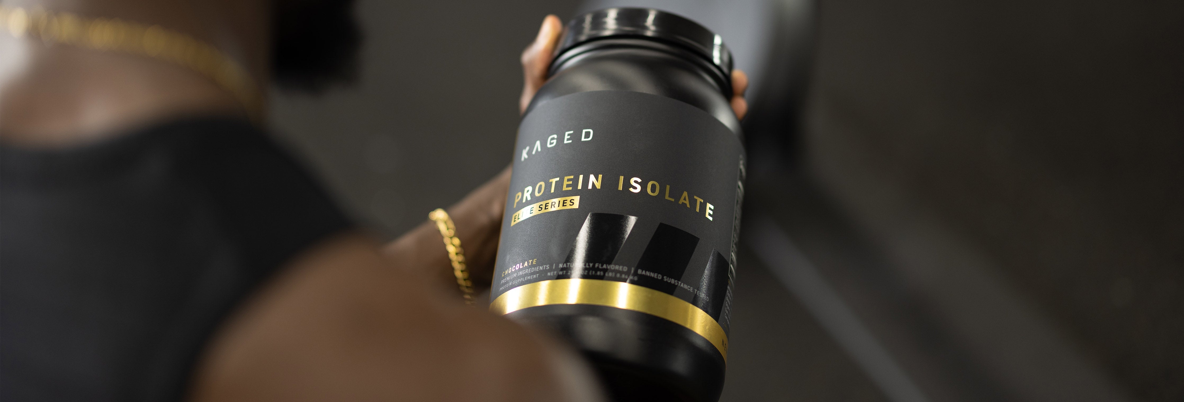 Close-up of KAGED Protein Isolate tub held in hand showing black label with gold trim and Protein Isolate text, blurred gym background