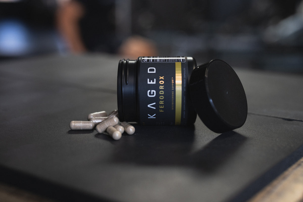Angled view of KAGED Ferodrox supplement jar on its side showcasing spilled LJ100 Tongkat Ali capsules on a dark gym mat