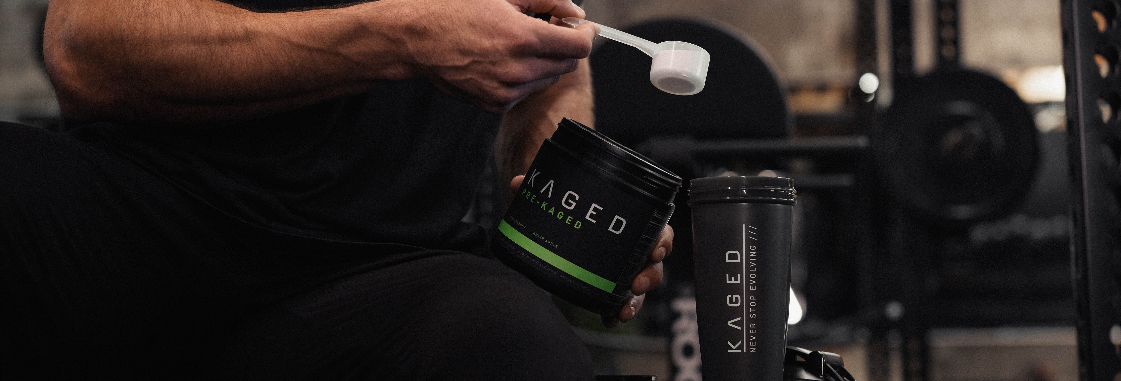 Person scooping Kaged powdered supplement into a Kaged shaker bottle at a gym