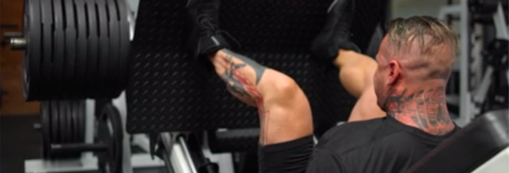 Rear view of a tattooed man performing a heavy leg press on a machine in a gym