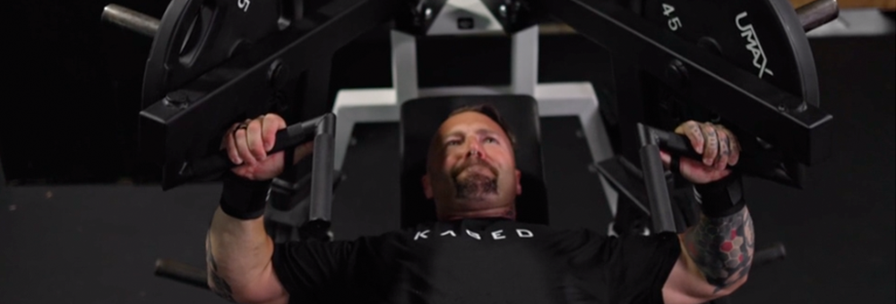 Man performing an incline chest press on a plate-loaded machine in a gym, wearing a black KAGED shirt