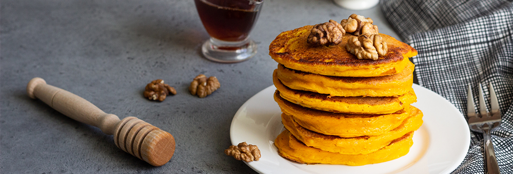 Stack of protein pancakes topped with walnuts on a white plate beside a honey dipper and fork on a gray countertop