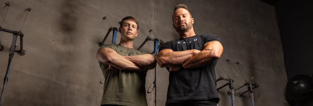 Two muscular men standing with arms crossed in a gym with concrete wall and suspension trainers in the background
