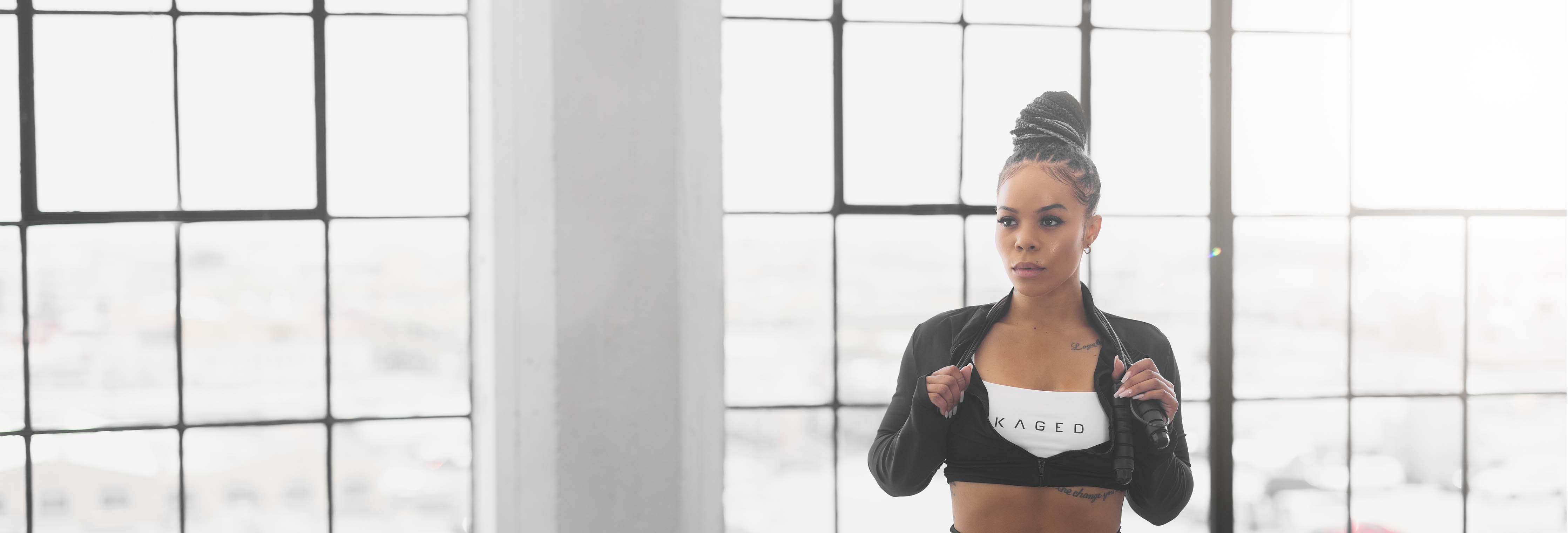 Woman adjusting a jacket in front of large industrial windows, wearing a white sports bra with KAGED logo