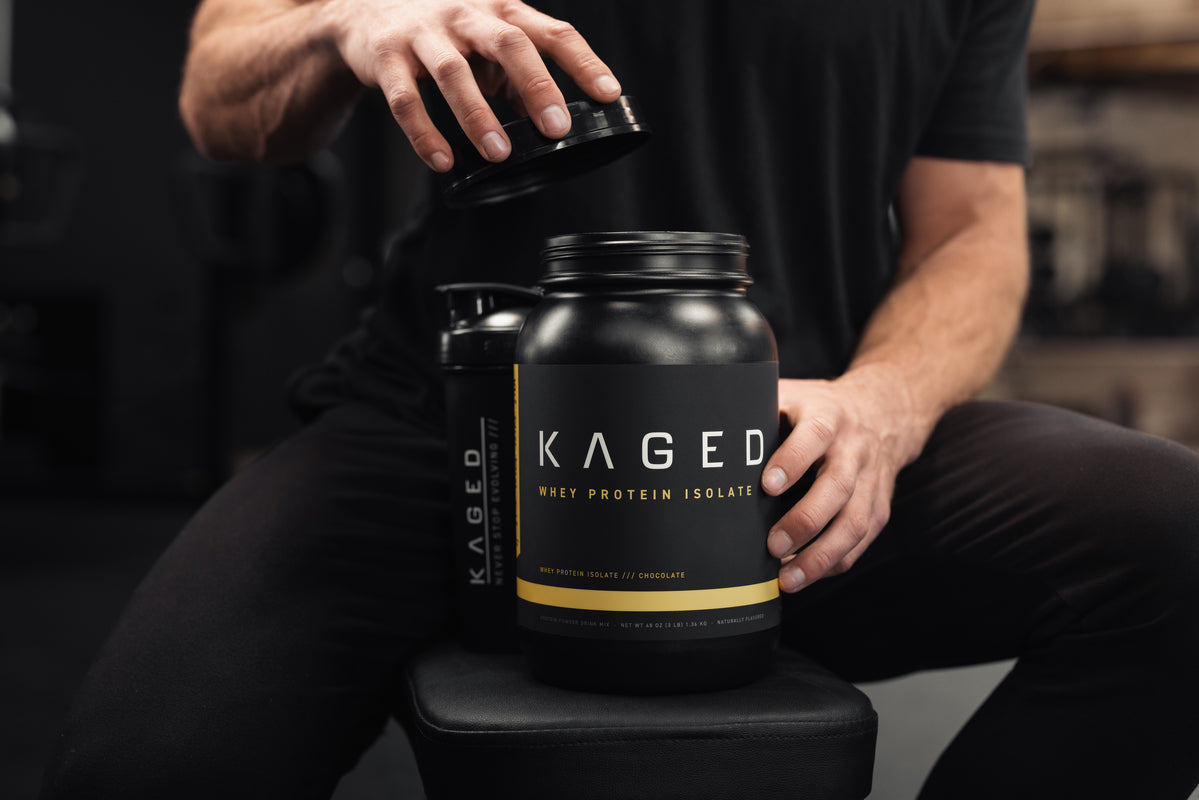 Front view of KAGED Whey Protein Isolate tub with lid lifted, label visible and shaker on a gym bench