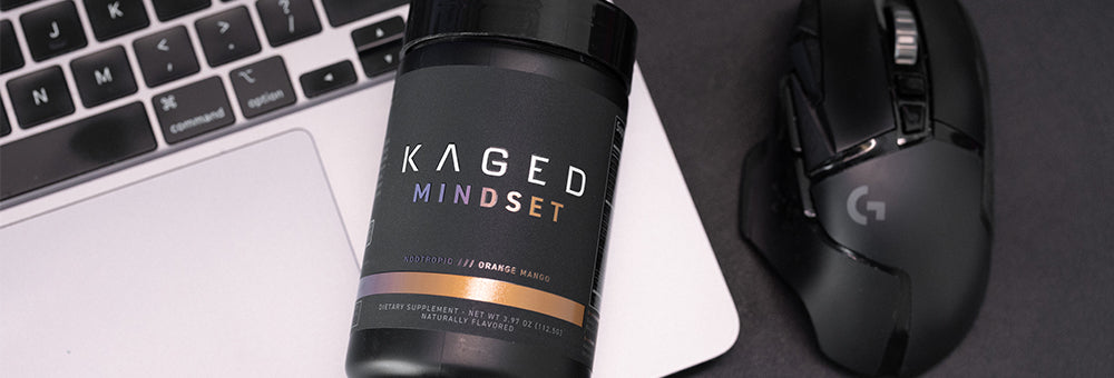 Angled view of KAGED Mindset nootropic supplement tub showing label and Orange Mango flavor next to laptop keyboard and mouse