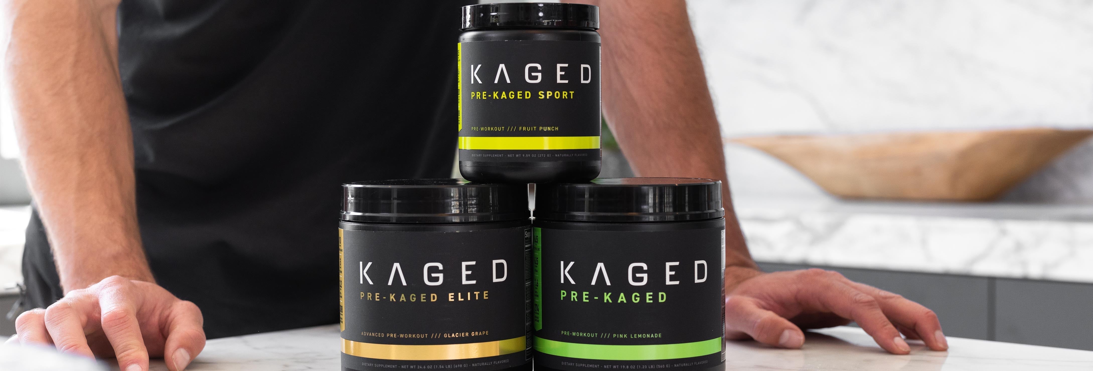 Front view of three KAGED pre-workout tubs showcasing colored label bands on marble countertop with hands behind