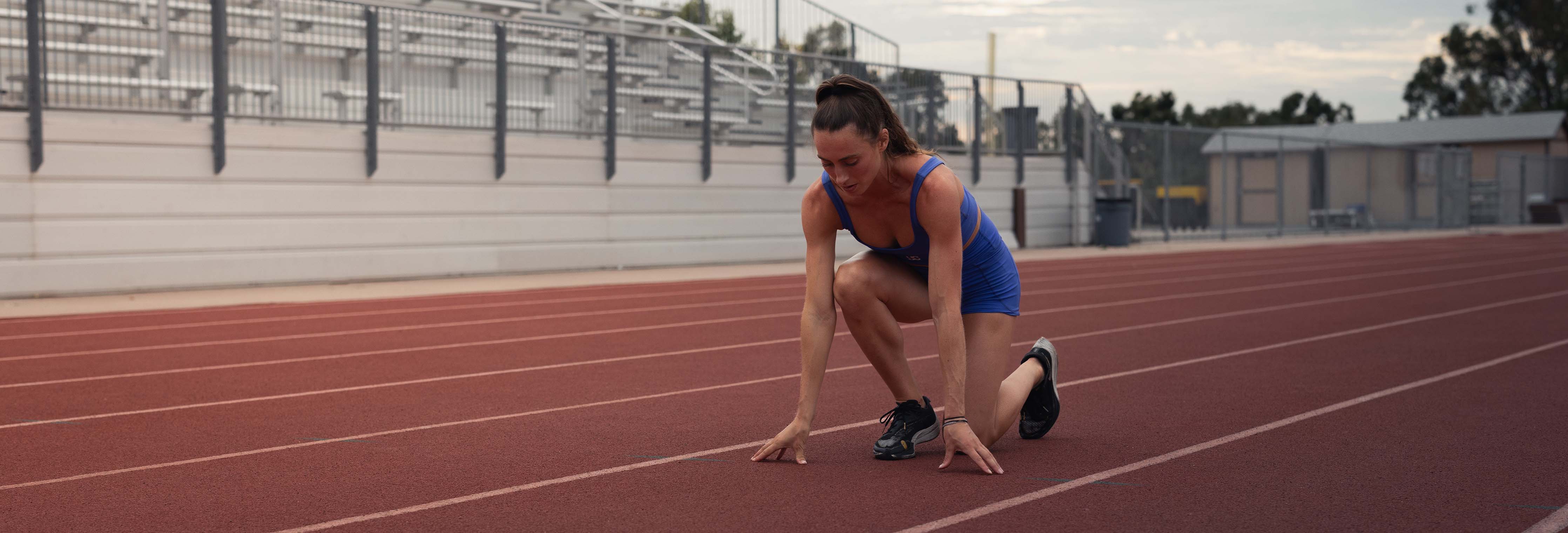 Woman runner in blue singlet kneeling in starting position on outdoor track at stadium