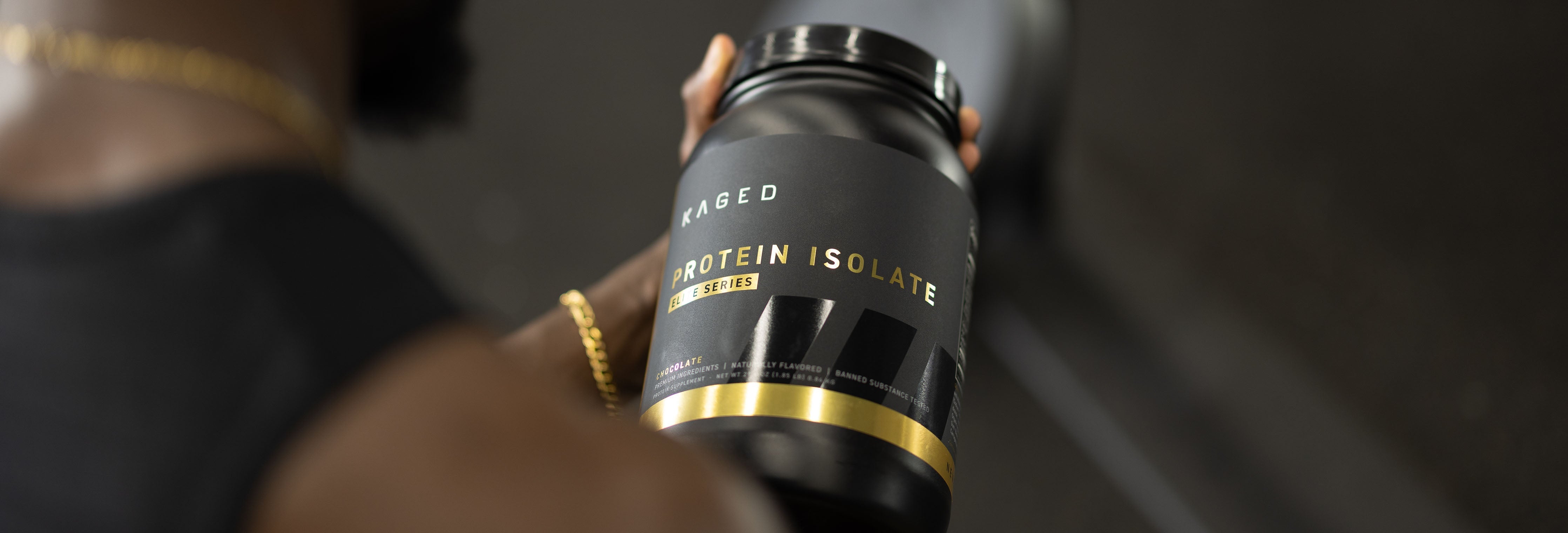 Angled close-up of KAGED Protein Isolate tub with black-and-gold label held by a person in a blurred gym background