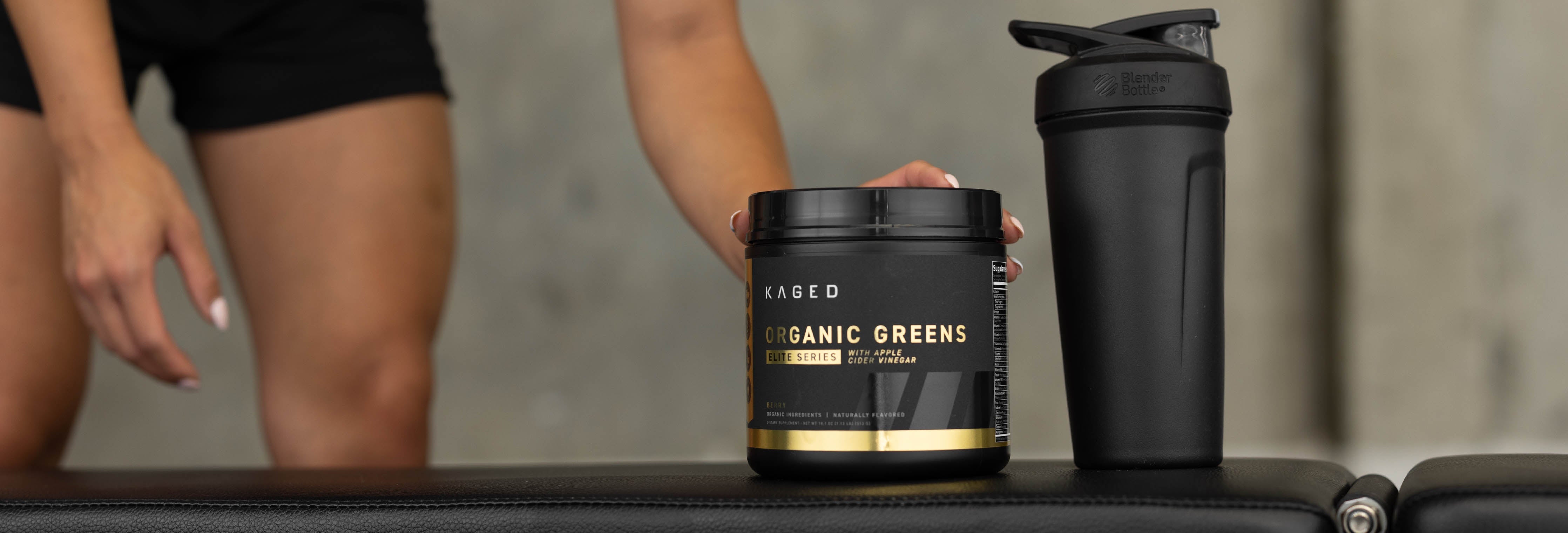 KAGED Organic Greens tub and black shaker on a gym bench with an athlete's legs visible