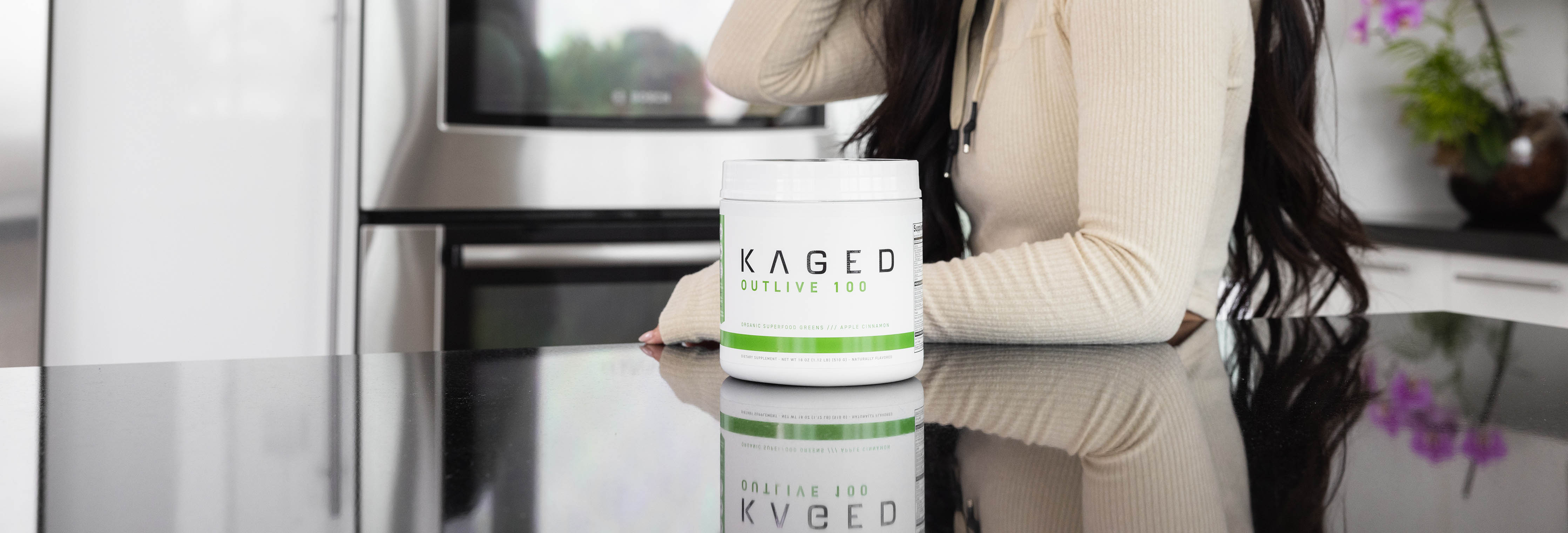 Front view of KAGED Outlive 100 white supplement tub with green label on a glossy black kitchen counter, woman leaning behind