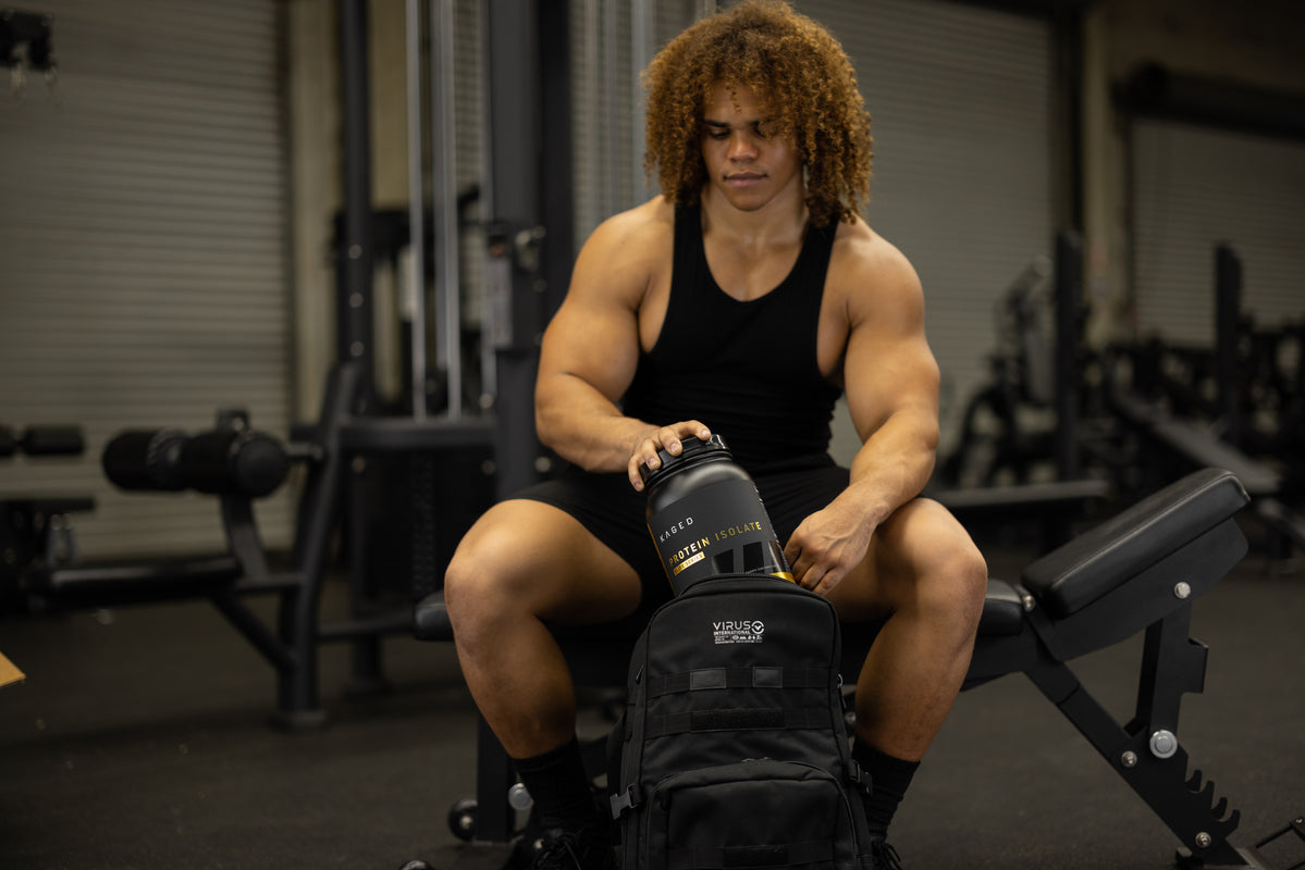 Athlete placing a KAGED Protein Isolate tub into a gym backpack while seated on a weight bench