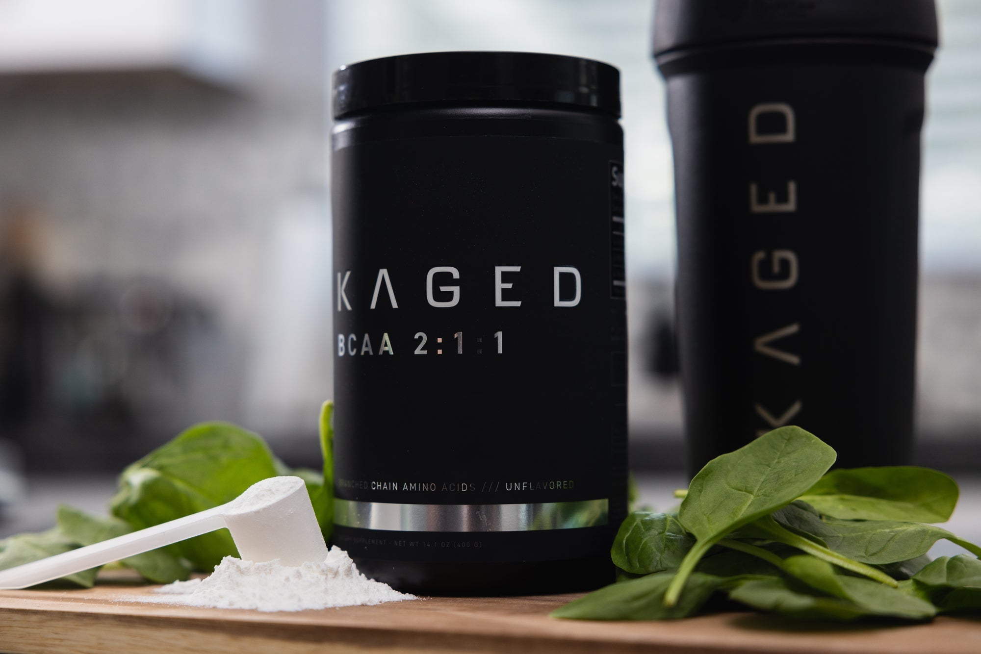 Front of KAGED BCAA 2:1:1 tub with scoop of powder, KAGED shaker, and spinach on a wooden board