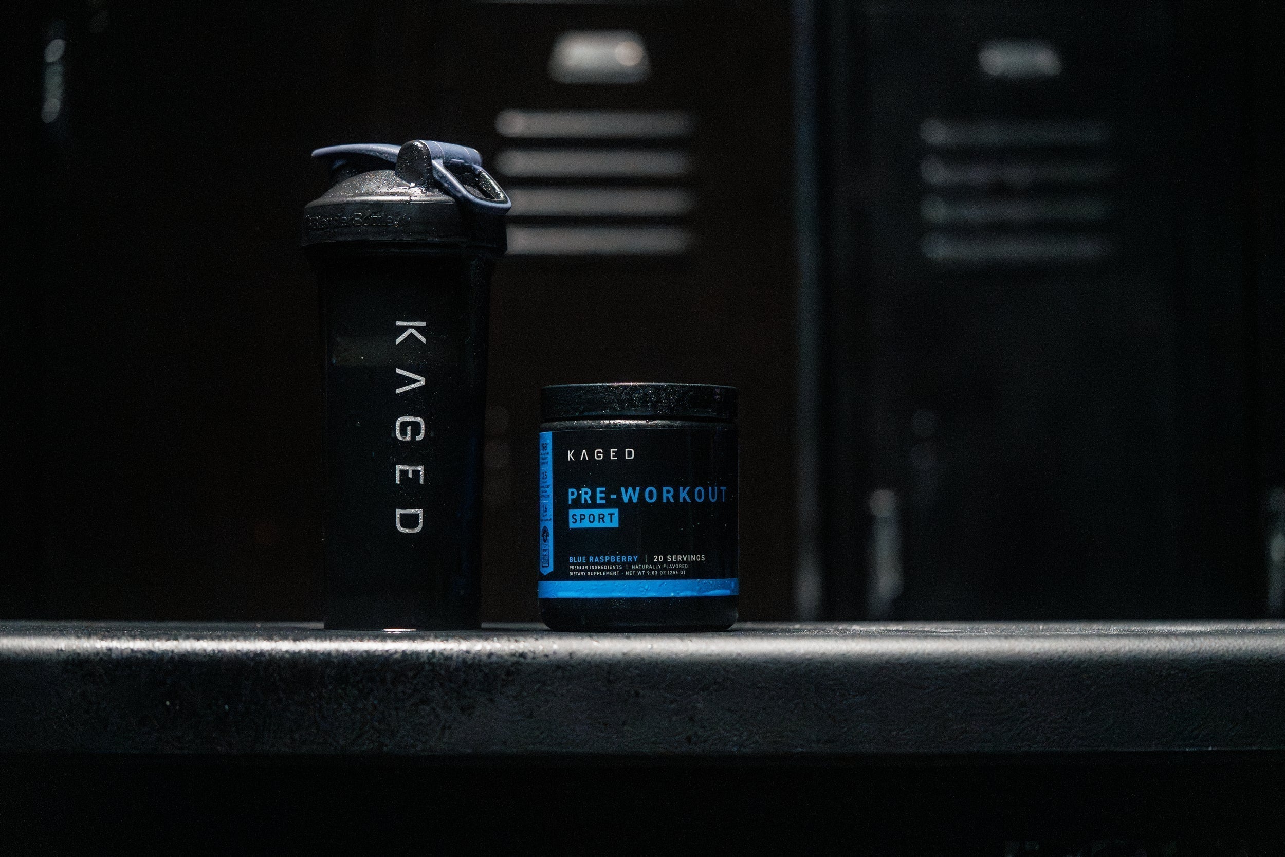 Front view of KAGED pre-workout tub and KAGED shaker on a gym bench, label reads PRE-WORKOUT SPORT against dark locker room