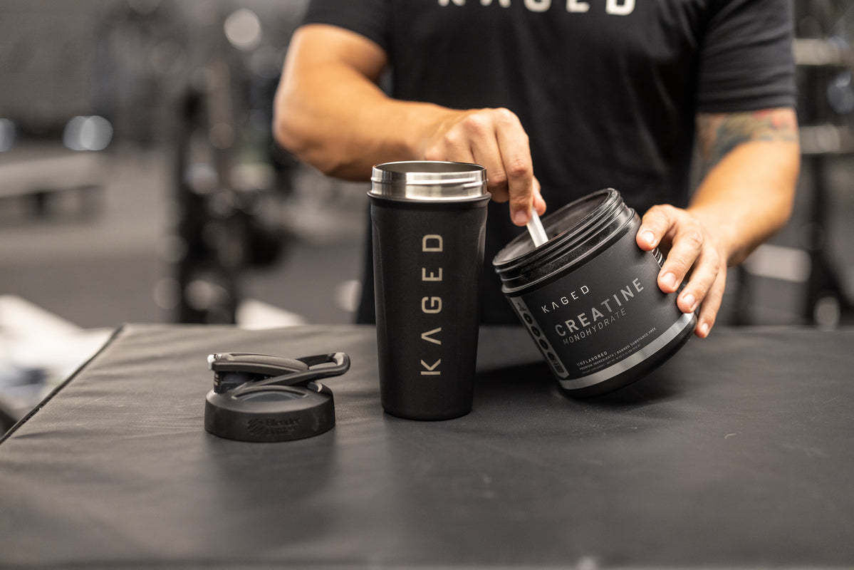 Front view of KAGED Creatine Monohydrate tub and black KAGED shaker cup on a gym bench, person scooping powder into the cup