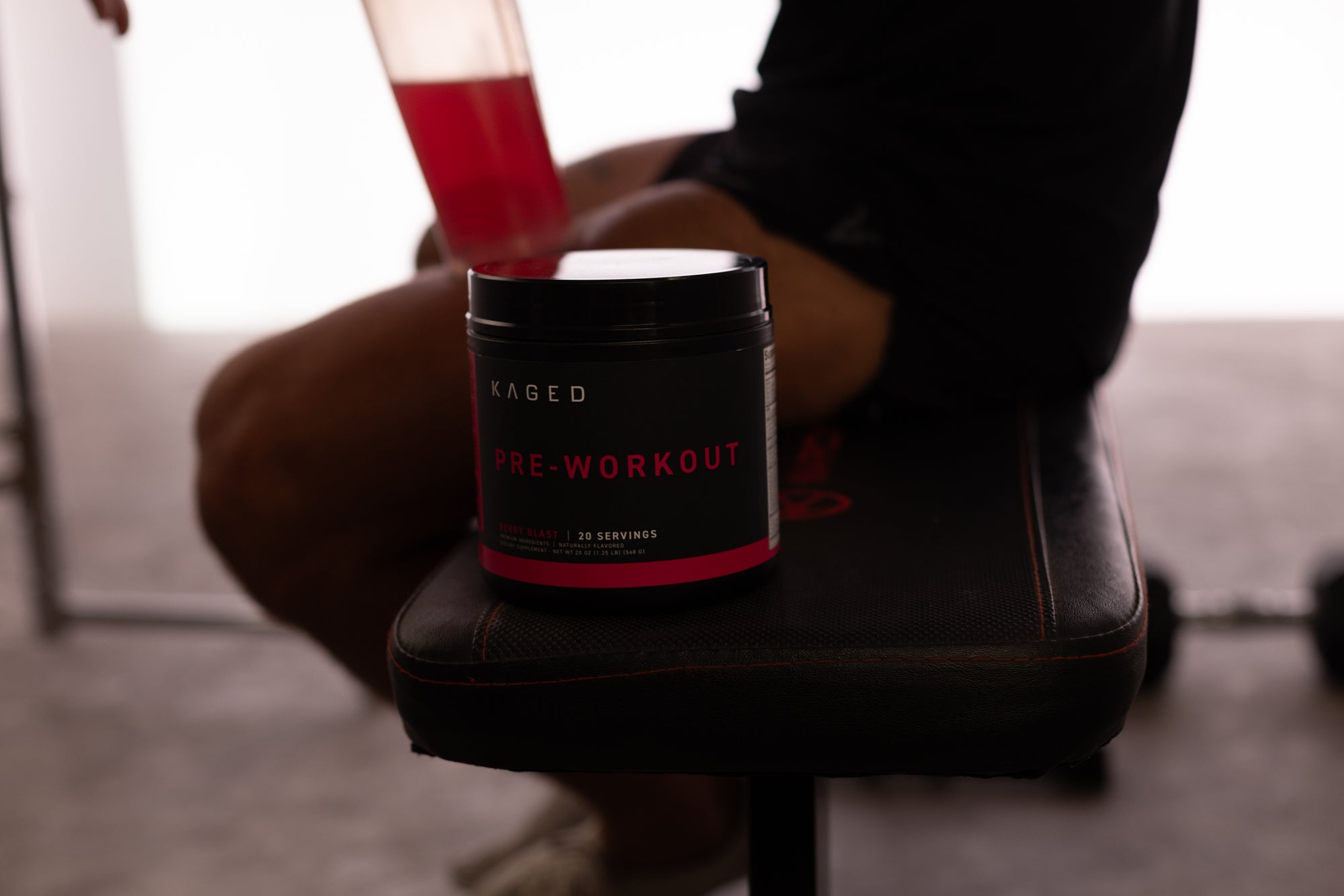 Front view of KAGED Pre-Workout tub showcasing black label and pink accent on a gym bench with an athlete holding a red shaker