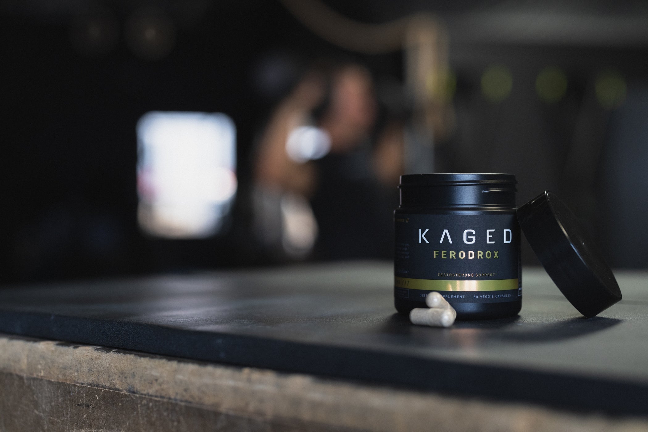 Front view of Kaged Ferodrox supplement jar showing label and two capsules on gym bench with blurred lifter in background