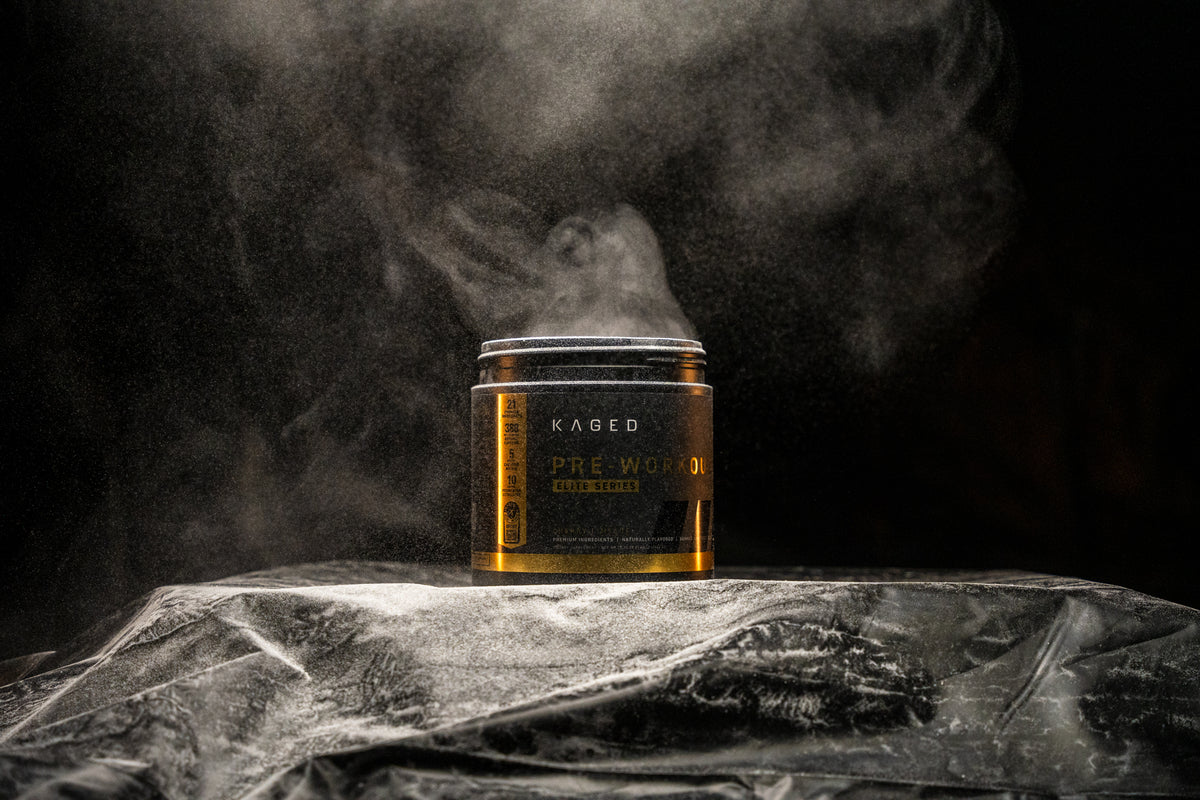 Front view of KAGED pre workout tub showing gold-accent label and powder mist rising against a dark backdrop