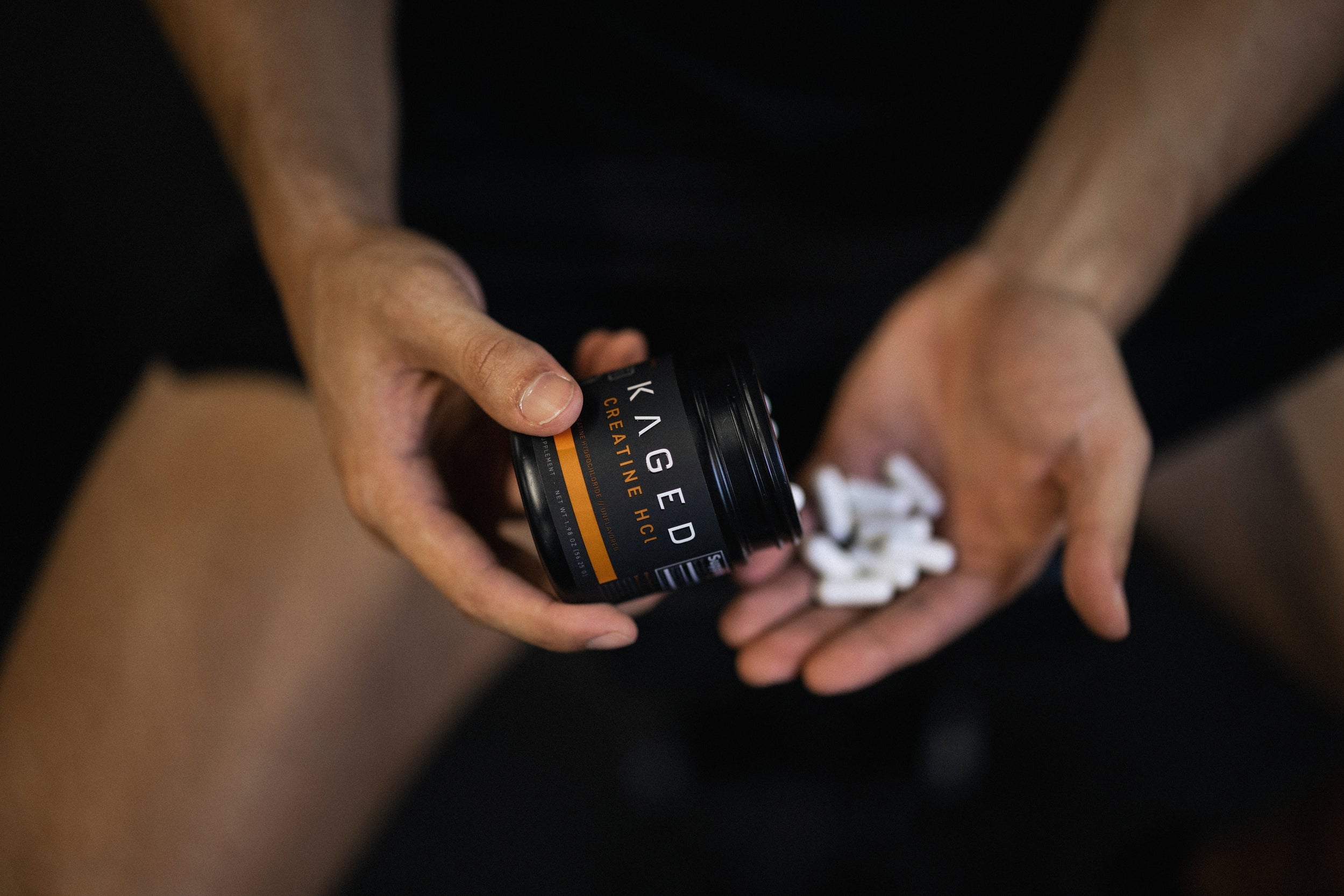Angled view of KAGED Creatine HCl jar pouring white capsules into a hand against a dark blurred background