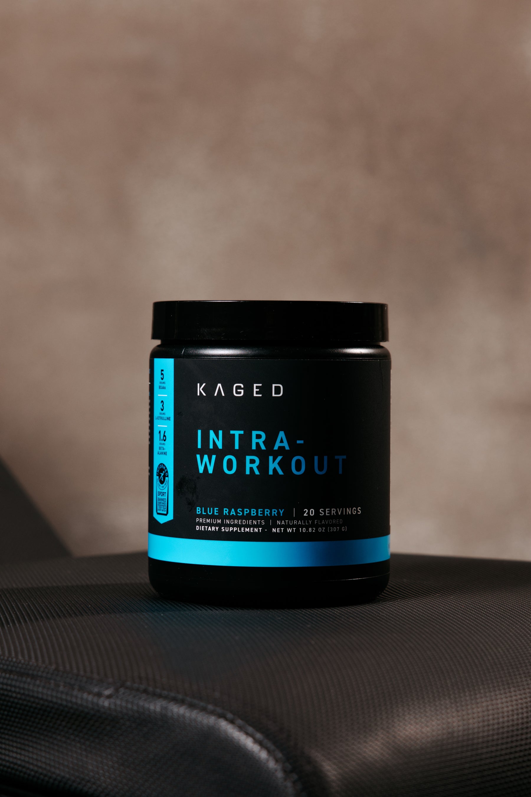 Front view of KAGED Intra-Workout supplement tub, Blue Raspberry flavor, 20 servings on a black container against a neutral brown backdrop