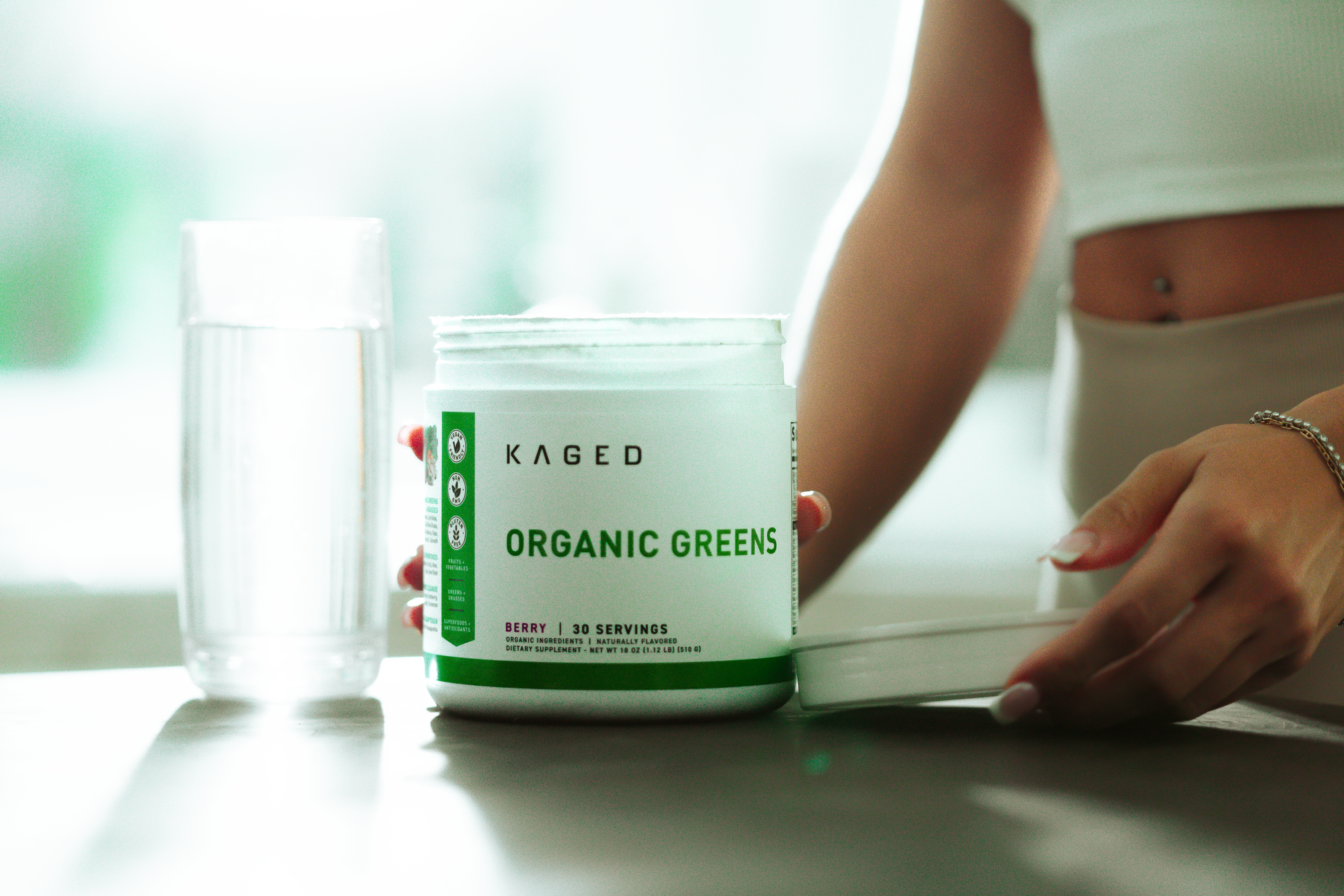 Front view of KAGED Organic Greens tub with lid off showing 'Organic Greens' label and 'Berry 30 servings' on a kitchen counter beside a glass of water