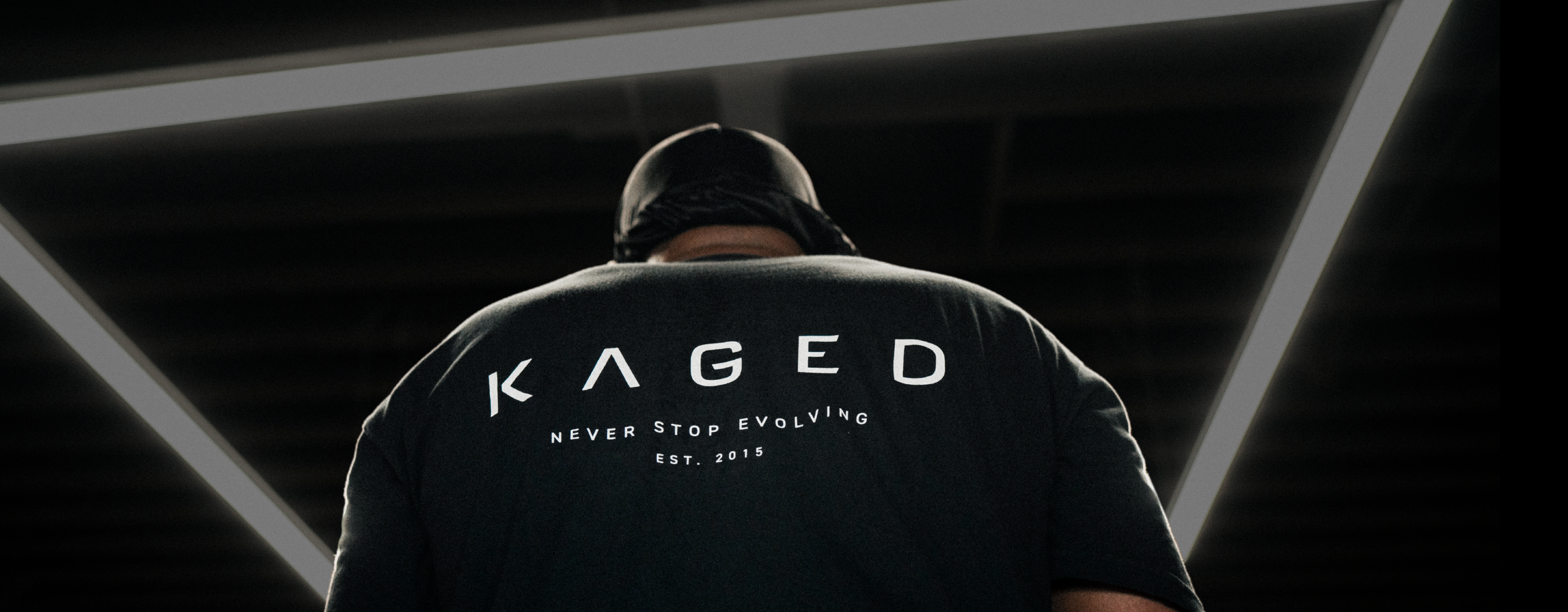 Person seen from behind wearing a black KAGED t-shirt reading KAGED Never Stop Evolving Est. 2015 under gym ceiling lights