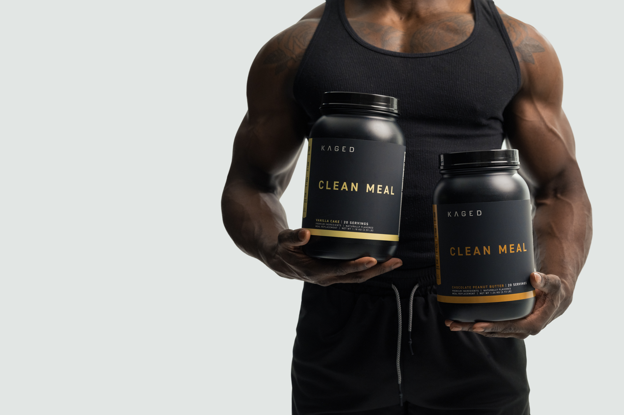 Two KAGED Clean Meal tubs (Vanilla Cake, Chocolate Peanut Butter) held by a man in a black tank top on pale gray background