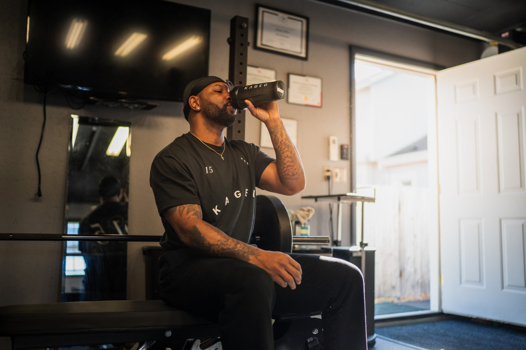 Man drinking a protein shake from a KAGED shaker while seated on a weight bench in a home gym