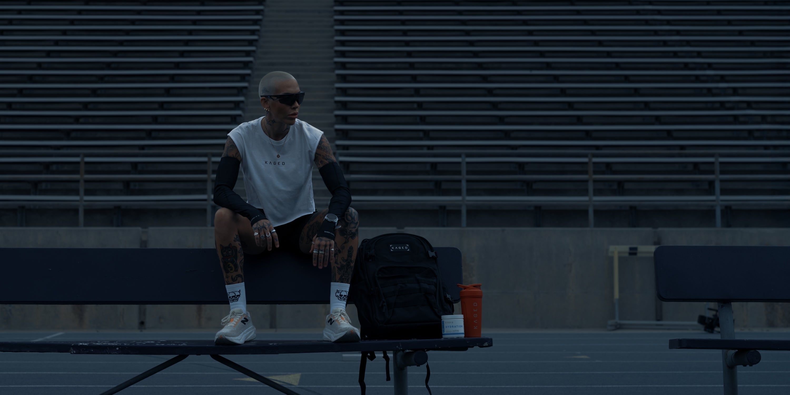 Athlete sitting on a stadium bench with a backpack, protein tub, and orange shaker bottle