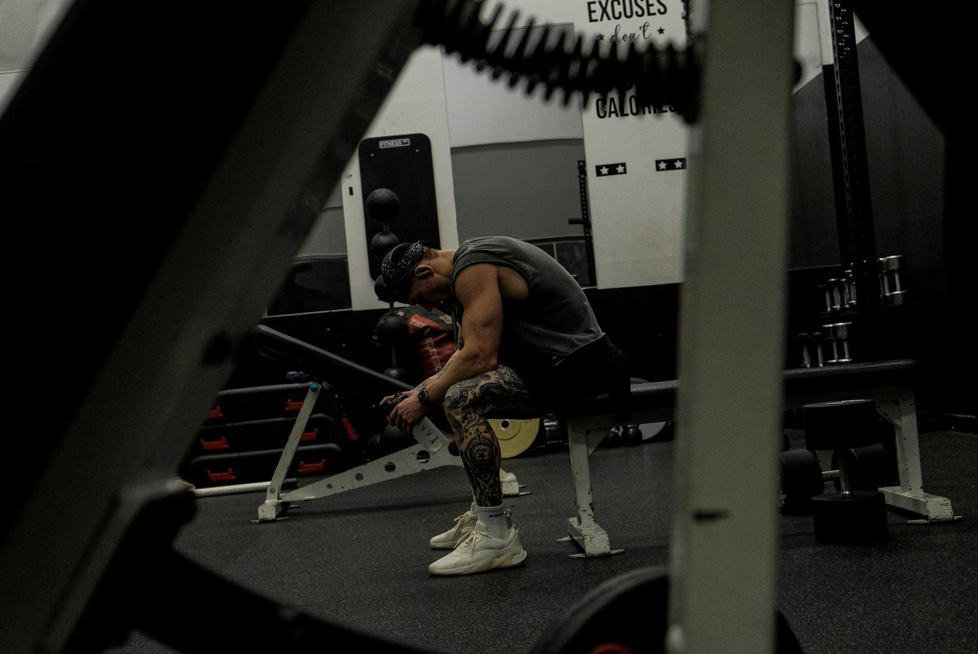 Tattooed man sitting on a gym bench resting and looking at a supplement shaker in a dimly lit weight room