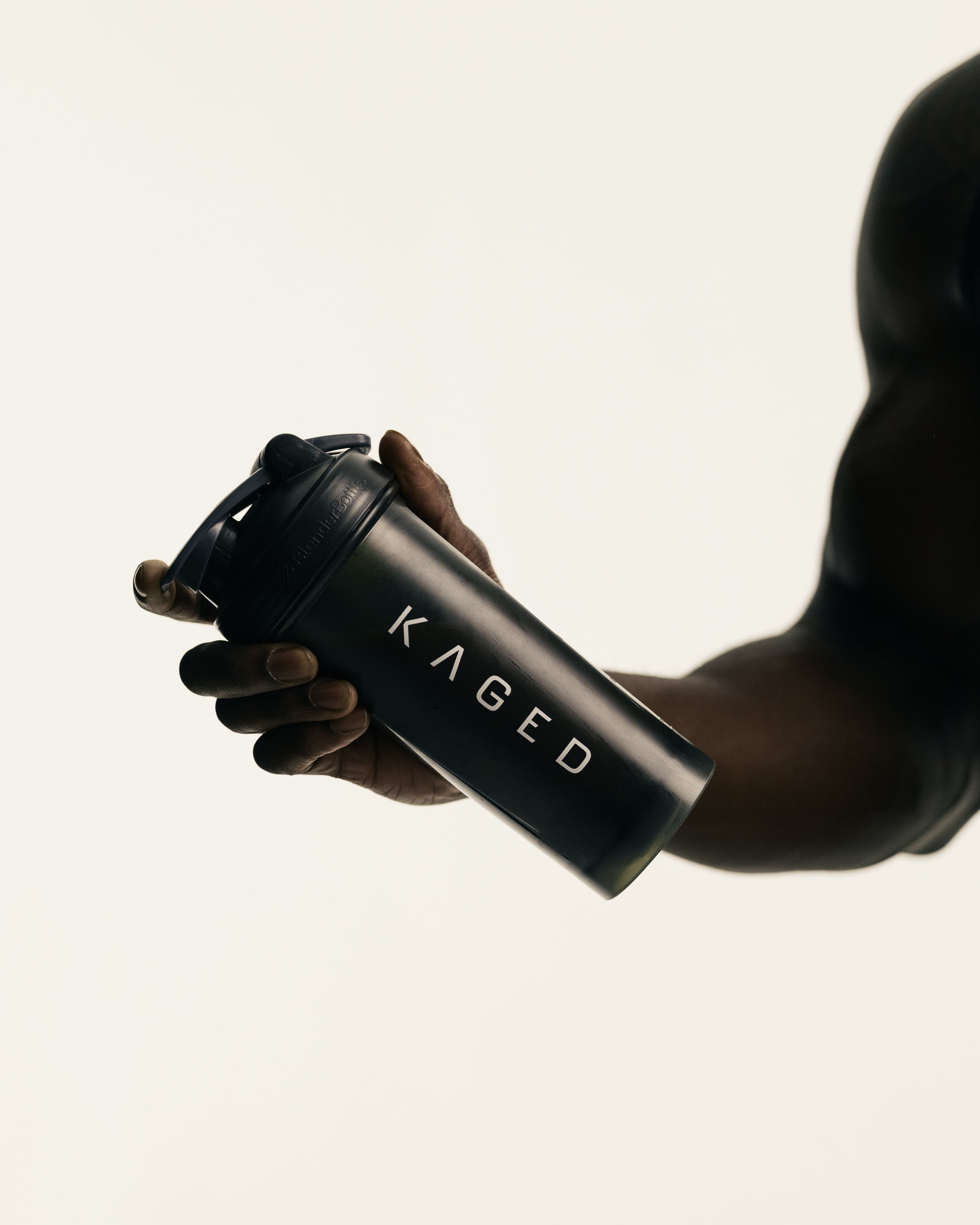 Handheld angled view of KAGED black shaker bottle showcasing KAGED logo and flip-top lid against a pale background