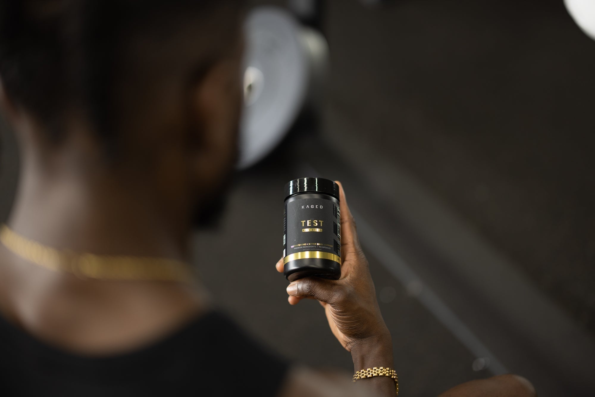 Close-up of KAGED TEST supplement jar held in hand, black-and-gold label reading TEST with blurred gym equipment in background