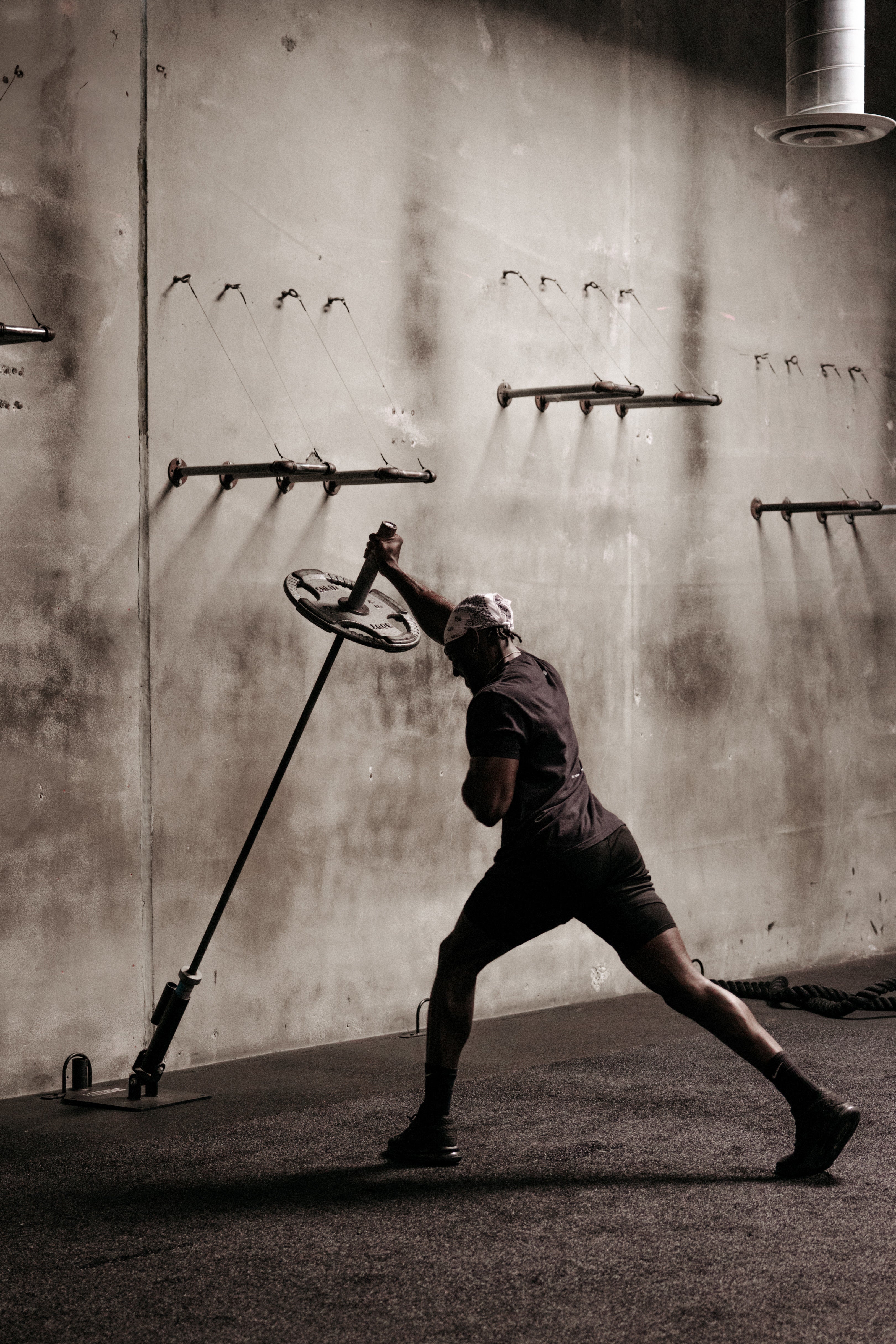 Man performing a landmine press in an industrial-style gym, lunging forward in a split stance against a concrete wall