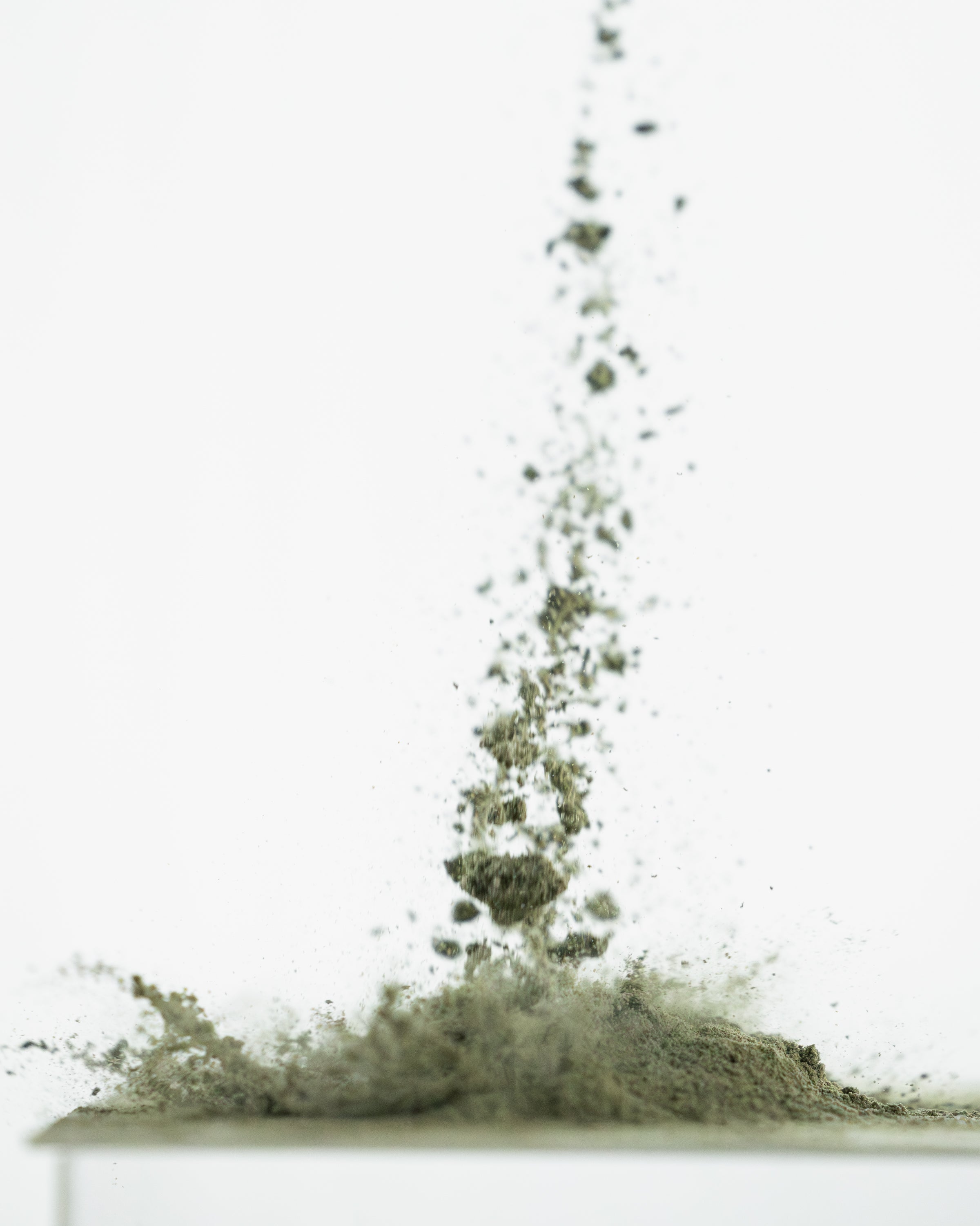 Close-up of greens supplement powder mid-air showcasing airborne fine green powder and clumps against a white background