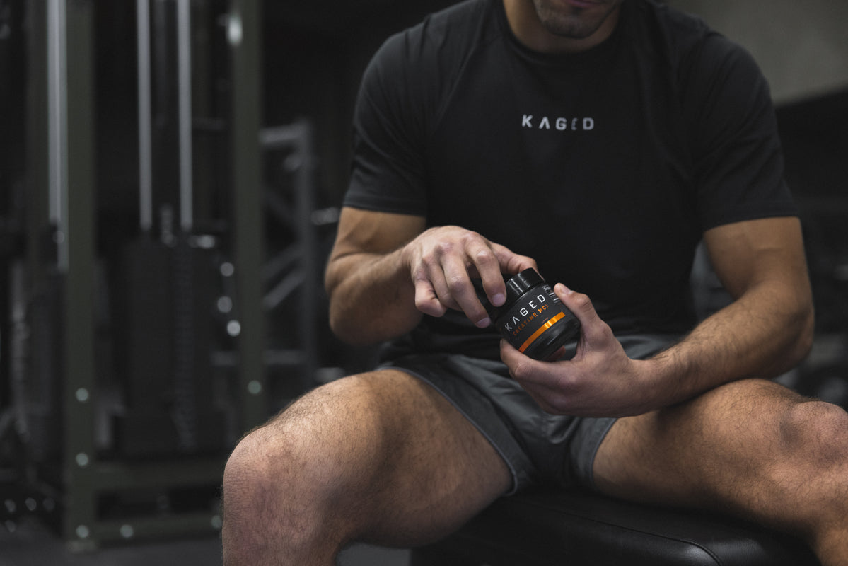 Athlete sitting on a gym bench holding a KAGED creatine container and checking the label, weight machines blurred behind him