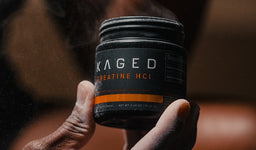 Close-up of a hand holding a Kaged Creatine HCL jar with powder mist in the air.