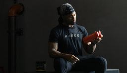 A person in a Kaged t-shirt sitting in a gym, holding a protein shake post-workout, looking relaxed and energized.
