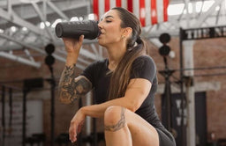 Athletic woman kneeling in a gym, drinking from a Kaged shaker bottle during a workout session.